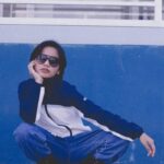 A woman in sunglasses and stylish outfit crouches against a blue wall, exuding coolness.
