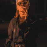 A retro-styled woman smokes while wearing vintage clothes and a camera outdoors.
