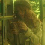 A woman with glasses and a beret enjoys reading in a warm, green-lit library environment.