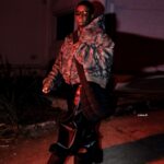 Person in stylish urban outfit at night, featuring camo jacket and sunglasses.