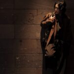 A fashionable person in a dark outfit leans against a textured wall, creating a moody urban scene.