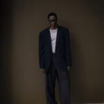 Stylish man wearing oversized suit poses in a modern, dimly-lit setting, exuding confidence.